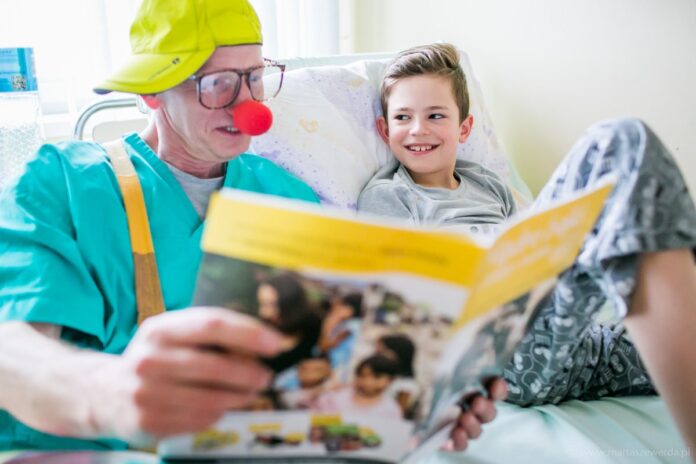 na zdjęciu znajduje się clown czytający choremu dziecku książkę